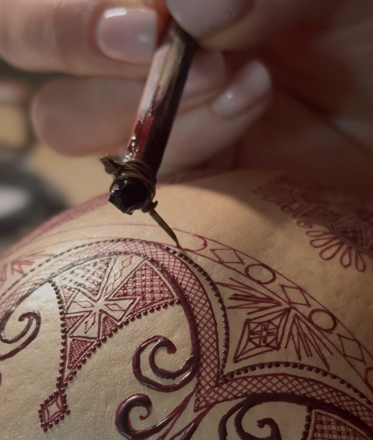 A hand using a traditional wax-writing tool (chișiță) to apply fine raised-wax ornaments to a natural eggshell