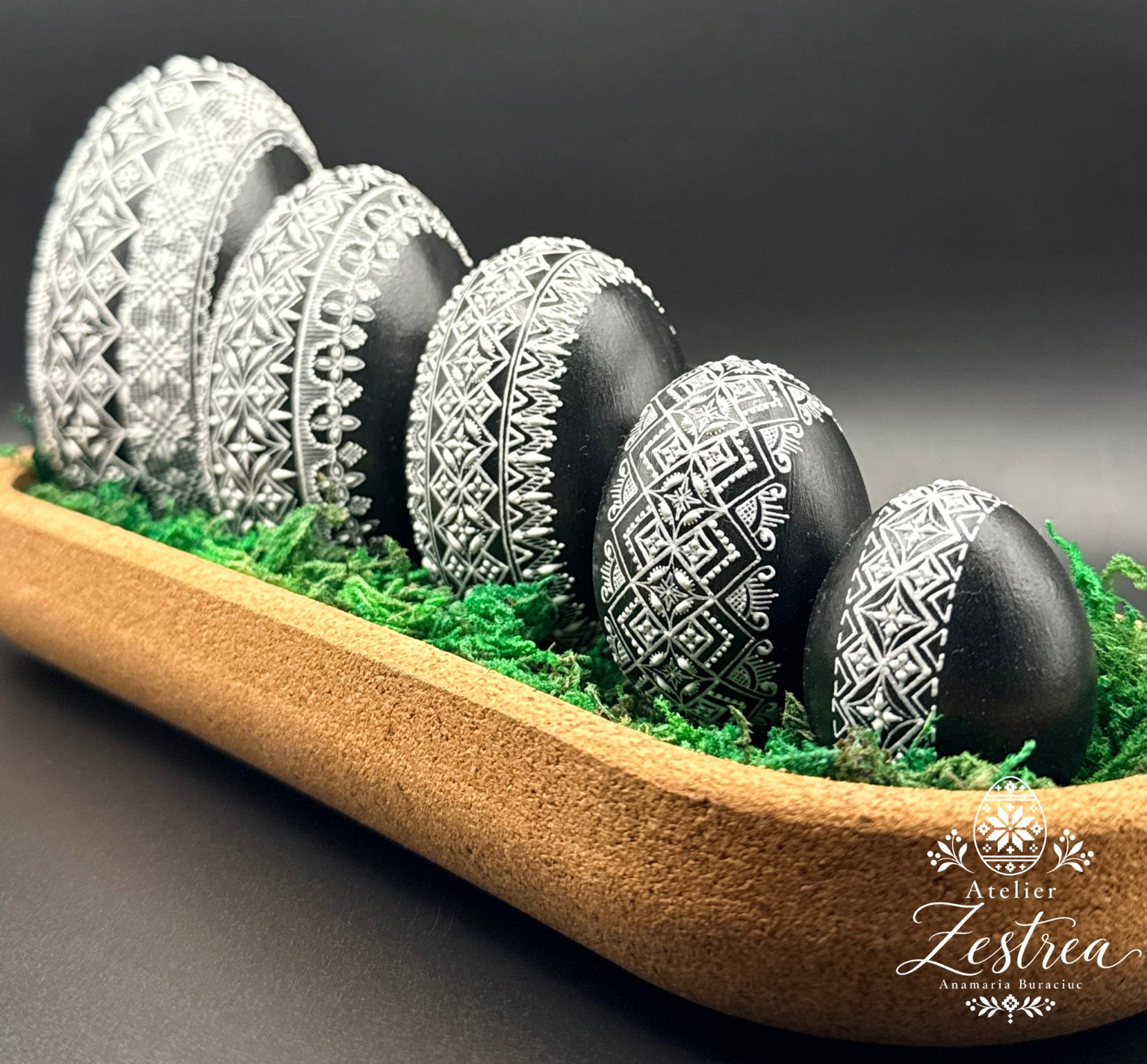 Black goose eggs with white geometric wax patterns, arranged on moss in a wooden tray